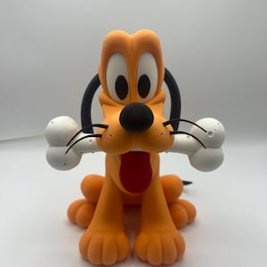Op de afbeelding: Een beeldje van Pluto, een cartoonhond, in een zittende positie. De oranje hond heeft een zwarte neus, oren en staart, met een rode tong die uitsteekt. Hij houdt een wit bot in zijn mond. Het beeldje staat op een wit oppervlak.