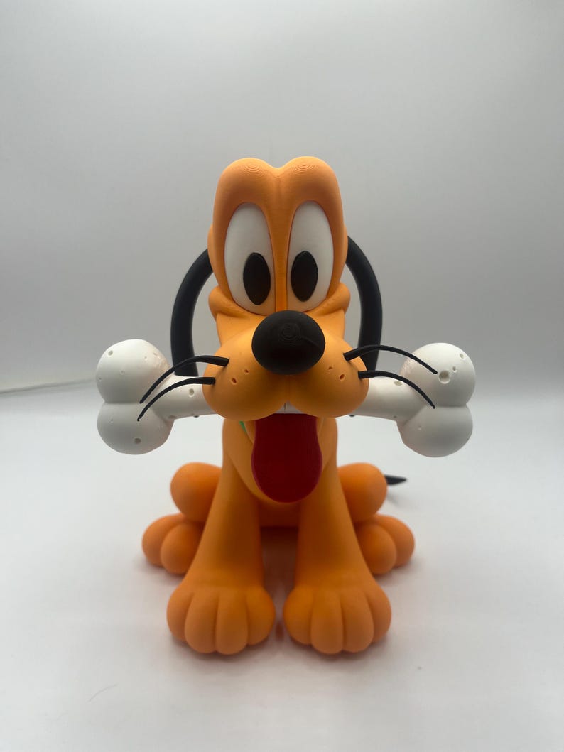 Op de afbeelding: Een beeldje van Pluto, een cartoonhond, in een zittende positie. De oranje hond heeft een zwarte neus, oren en staart, met een rode tong die uitsteekt. Hij houdt een wit bot in zijn mond. Het beeldje staat op een wit oppervlak.
