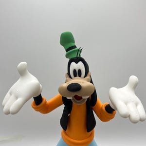 Op de afbeelding: Een beeldje van Goofy, een Disney-stripfiguur, met een oranje shirt, een blauwe broek en bruine schoenen. Hij heeft witte handschoenen en een groene hoed. Het beeldje staat met uitgestrekte armen.