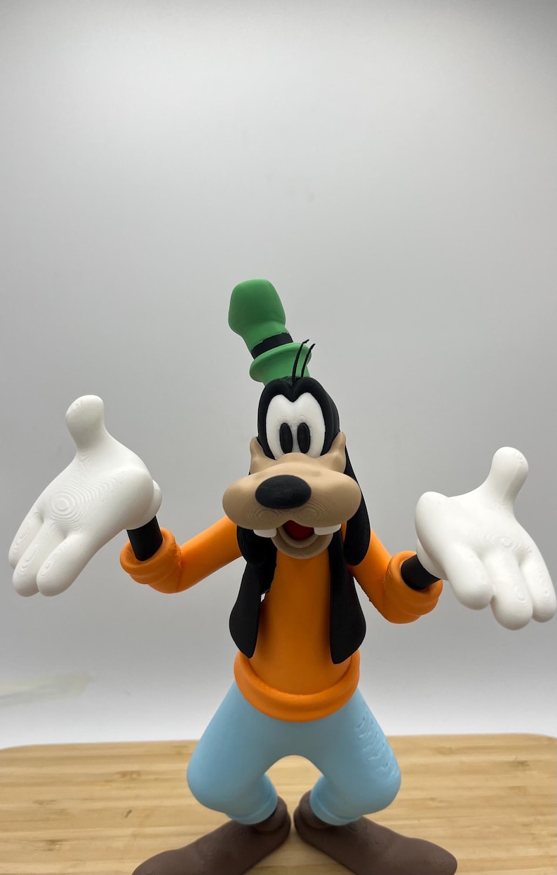 Op de afbeelding: Een beeldje van Goofy, een Disney-stripfiguur, met een oranje shirt, een blauwe broek en bruine schoenen. Hij heeft witte handschoenen en een groene hoed. Het beeldje staat met uitgestrekte armen.