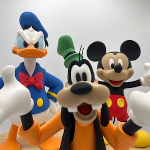 Op de afbeelding: Beeldjes van Disney-personages Donald Duck, Goofy en Mickey Mouse. Donald Duck draagt een blauw matrozenpak met een rode vlinderdas. Goofy draagt een oranje outfit en een groene hoed. Mickey Mouse draagt een rode broek en witte handschoenen.