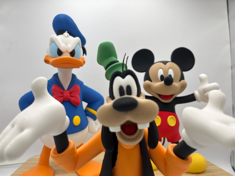 Op de afbeelding: Beeldjes van Disney-personages Donald Duck, Goofy en Mickey Mouse. Donald Duck draagt een blauw matrozenpak met een rode vlinderdas. Goofy draagt een oranje outfit en een groene hoed. Mickey Mouse draagt een rode broek en witte handschoenen.