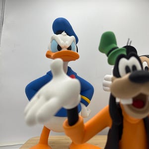 Op de afbeelding: Close-up van beeldjes van Donald Duck en Goofy. Donald Duck draagt een blauw matrozenhemd en hoed, met een oranje snavel en voeten. Goofy draagt een oranje shirt, zwart vest en groene hoed. De beeldjes staan op een houten oppervlak.