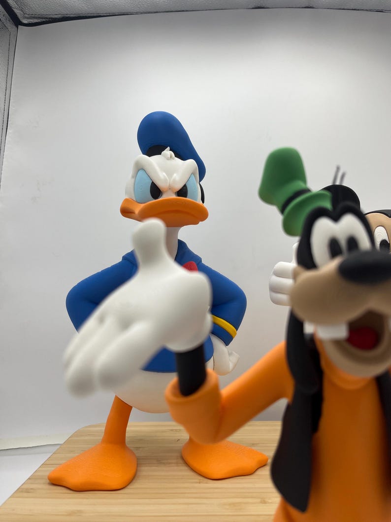Op de afbeelding: Close-up van beeldjes van Donald Duck en Goofy. Donald Duck draagt een blauw matrozenhemd en hoed, met een oranje snavel en voeten. Goofy draagt een oranje shirt, zwart vest en groene hoed. De beeldjes staan op een houten oppervlak.