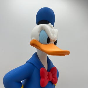 Op de afbeelding: Een 3D-geprinte Donald Duck-beeldje. Het heeft een blauw matrozenpak met gele details, een rode vlinderdas en een blauwe hoed. De eend heeft een oranje snavel, een wit gezicht en blauwe ogen, trouw aan het personage.