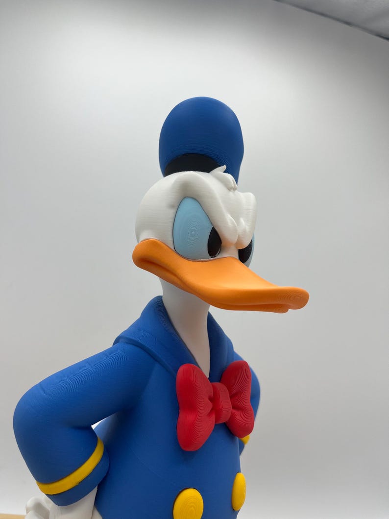 Op de afbeelding: Een 3D-geprinte Donald Duck-beeldje. Het heeft een blauw matrozenpak met gele details, een rode vlinderdas en een blauwe hoed. De eend heeft een oranje snavel, een wit gezicht en blauwe ogen, trouw aan het personage.