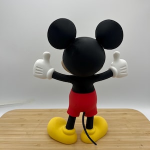 Op de afbeelding: Een Mickey Mouse-beeldje, van achteren gezien, met armen omhoog in een duim omhoog gebaar. De figuur heeft een zwarte kop met grote oren, een zwart lichaam, rode shorts, witte handschoenen en gele schoenen. Het beeldje staat op een houten oppervlak.