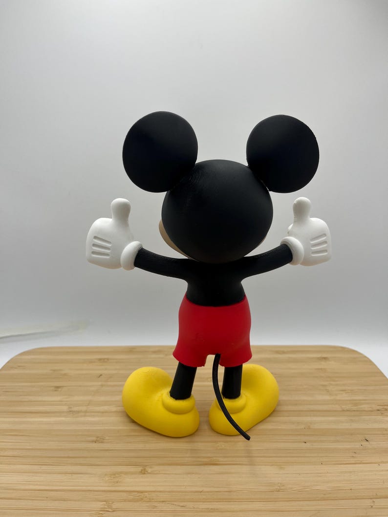 Op de afbeelding: Een Mickey Mouse-beeldje, van achteren gezien, met armen omhoog in een duim omhoog gebaar. De figuur heeft een zwarte kop met grote oren, een zwart lichaam, rode shorts, witte handschoenen en gele schoenen. Het beeldje staat op een houten oppervlak.