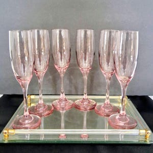 May include: A set of six tall, slender pink glass champagne flutes with smooth, rounded bowls and clear stems, arranged in two rows on a rectangular mirrored serving tray featuring a clear glass handle and gold-toned metal accents at the corners.