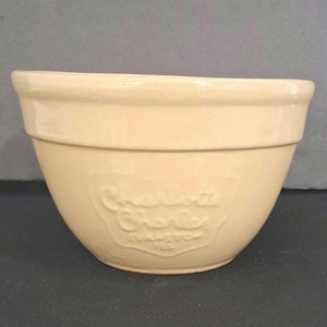 May include: A cream-colored ceramic mixing bowl with a smooth, glossy finish and a wide, flared rim. The front of the bowl features an embossed, shield-shaped logo with the text Charlotte Charles Evanston, Ill. molded into the surface.