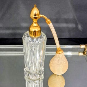 May include: A vintage perfume bottle with a clear, textured glass body and a gold-colored atomizer. The atomizer has a bulbous, beige fabric puff and a gold-colored nozzle. The bottle is displayed on a reflective surface.