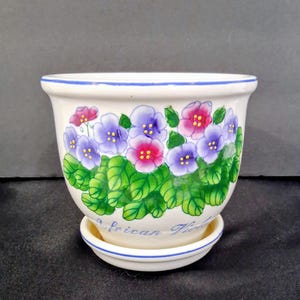 May include: Cream-colored ceramic flower pot with a matching saucer. The pot is decorated with a floral design featuring purple, pink, and green flowers and leaves. The rim has a blue trim, and the words "African Violets" are written on the pot.