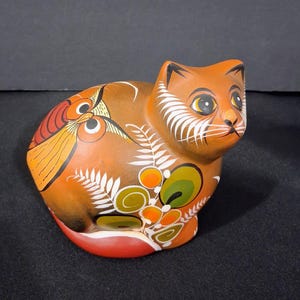 May include: A ceramic cat figurine in shades of brown, orange, and white. The cat is decorated with floral and owl-like patterns. The cat's eyes are yellow, and it has white whiskers. The figurine is sitting with its tail curled.