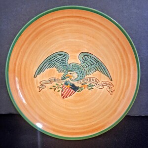 May include: A round ceramic plate with a warm orange, swirled background and a dark green rim. The center features a raised, embossed design of a bald eagle with spread wings, holding a banner that reads E PLURIBUS UNUM above a shield.