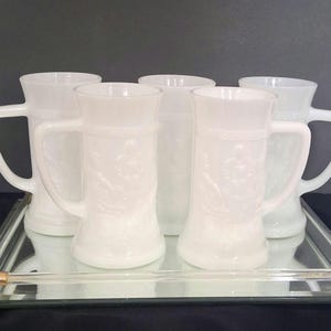 May include: A set of five white milk glass beer steins featuring embossed figural designs on the exterior. Each stein has a sturdy handle and a flared rim. The collection is arranged on a rectangular mirrored tray with a clear glass rail.