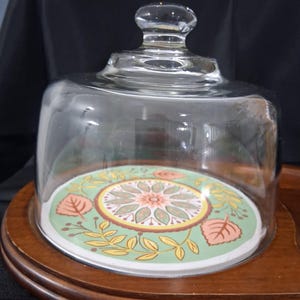 May include: A clear glass cake dome with a round, decorative base. The base features a floral design in shades of green, orange, and yellow. The dome sits on a wooden, tiered base, creating a vintage aesthetic. The dome has a clear glass knob.