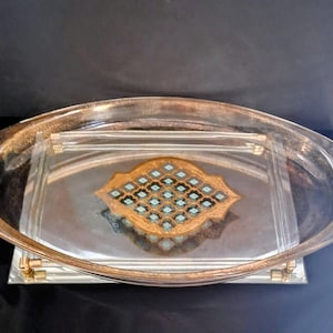May include: An oval-shaped serving tray with a clear glass rim and a decorative gold and silver interior. The tray features a central design with blue and gold accents. The tray has gold-colored handles.