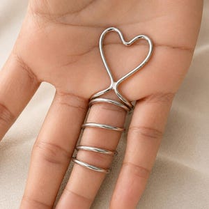 May include: A silver-toned metal finger splint featuring a heart-shaped design at the top. The structure consists of a continuous wire that wraps around the finger in four distinct horizontal coils, providing a secure fit along the digit.