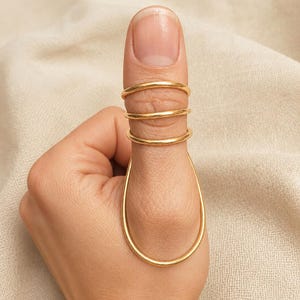 May include: A gold-colored metal splint ring featuring three thin, circular bands that wrap around the upper portion of the thumb, connected to a larger, elongated oval-shaped loop that encircles the base of the thumb joint.