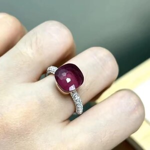 Könnte beinhalten: Ring mit einem quadratisch geschliffenen, tiefroten Edelstein. Der Silberring ist mit kleineren, klaren Steinen besetzt. Der Ring wird an einem Finger gezeigt.