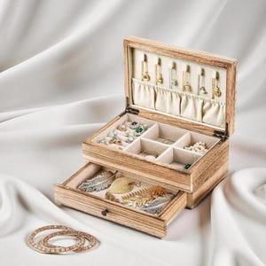 May include: A wooden jewelry box with multiple compartments and a pull-out drawer. The box is open, revealing various pieces of jewelry, including rings, necklaces, and earrings. The drawer contains decorative feather-shaped ornaments. A set of gold bangles is in the foreground.