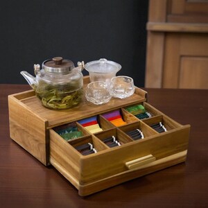 Puede incluir: Una caja de té de madera con un cajón extraíble, que contiene bolsitas de té. Una tetera de vidrio con hojas de té, un cuenco de vidrio pequeño y dos vasos pequeños están encima. La caja tiene un asa de latón.