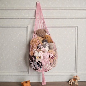 May include: A pink macrame hanging storage net filled with various plush toys, including teddy bears and dog-shaped stuffed animals. The net is suspended from a wall, offering a decorative and functional storage solution for children's toys.