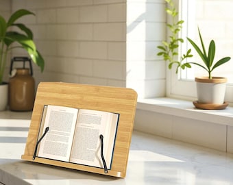 May include: A light-colored bamboo book stand with a book open to a page of text. The stand has a sloped design and black metal page holders. The stand is on a white countertop near a window with plants.