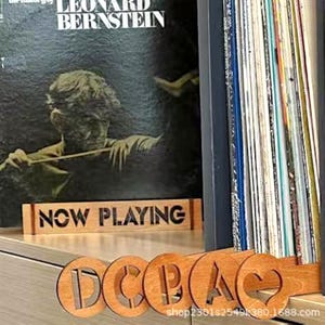 May include: A wooden "Now Playing" sign and a record divider with the letters "DCBAC" cut out. The background features a stack of vinyl records and a record album cover with the text "LEONARD BERNSTEIN".