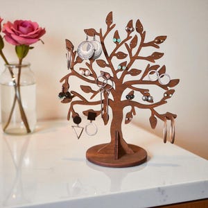 Può includere: Un albero portagioielli in legno a forma di albero con foglie, che contiene vari orecchini. L'albero è marrone e poggia su una base rotonda. Un vaso con una rosa rosa è sullo sfondo.