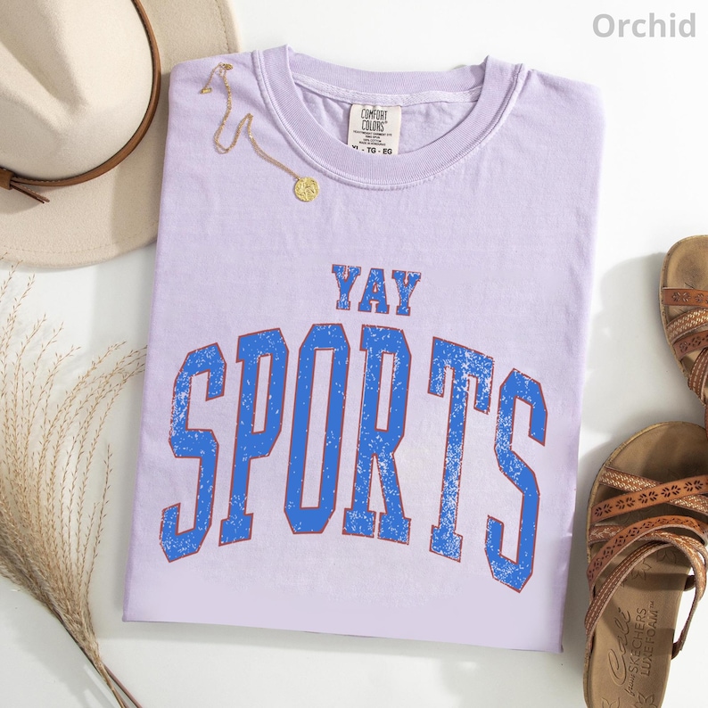 a purple t-shirt with the text "YAY SPORTS" printed on it, along with a beige hat and sandals.