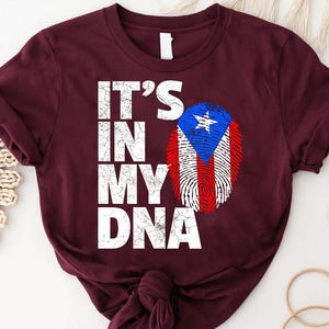 Puede incluir: Camiseta burdeos con la frase "IT'S IN MY DNA" en letras blancas. Un diseño de huella dactilar con los colores de la bandera de Puerto Rico está en el lado derecho. La camiseta es de material suave.