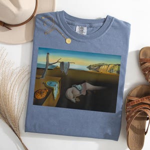 May include: A blue Comfort Colors t-shirt featuring a surrealist painting with melting clocks, a coastal landscape, and a tree branch. A gold necklace and a beige hat are also visible.