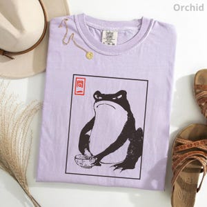 May include: A light purple t-shirt featuring a black and white graphic of a frog holding a bowl. The graphic is enclosed in a black rectangular frame with red Japanese characters in the top left corner. The shirt has a crew neckline and a tag.