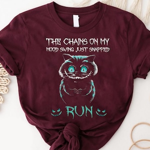 Op de afbeelding: Bordeauxrood t-shirt met een afbeelding van een Cheshire Cat met turquoise ogen en accenten. De tekst "THE CHAINS ON MY MOOD SWING JUST SNAPPED" staat boven de kat en "RUN" eronder. Het ontwerp is in wit en turquoise.