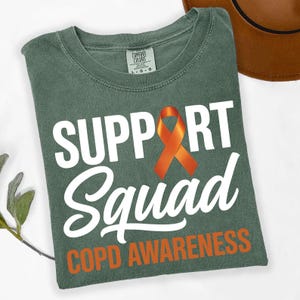 Puede incluir: Camiseta verde con las palabras "SUPPORT Squad" en blanco, un lazo naranja y "COPD AWARENESS" en naranja. La camiseta es de un material suave, probablemente de ajuste cómodo.