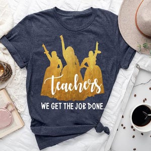 May include: Dark blue t-shirt featuring a gold silhouette of three figures with the word "Teachers" in white script. Below the word is the phrase "WE GET THE JOB DONE." The shirt is knotted at the bottom.