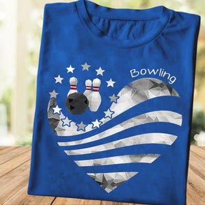 May include: Blue t-shirt with a bowling-themed design. The graphic features a black bowling ball, two white bowling pins with red stripes, and a heart-shaped design with gray and white stripes and stars. The word "Bowling" is written in white.