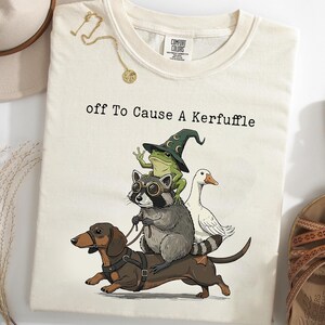 May include: A cream-colored t-shirt featuring a graphic of a dachshund wearing a harness, a raccoon with goggles, a frog in a wizard hat, and a white duck. The text "off To Cause A Kerfuffle" is printed above the animals in a black typewriter font.
