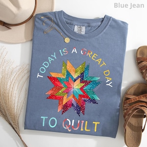 May include: A blue jean colored t-shirt featuring a multi-colored geometric star pattern in the center. The text "TODAY IS A GREAT DAY TO QUILT" is printed in a circular arc around the star design. The shirt has a crew neckline and a tag.