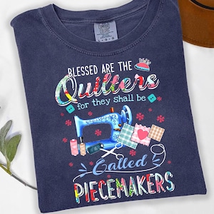 May include: Navy blue t-shirt with the text "Blessed are the Quilters for they shall be called PIECEMAKERS." The design includes a blue sewing machine, spools of thread, and fabric squares. The shirt is a Comfort Colors brand.