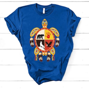 May include: Royal blue t-shirt featuring a turtle design with a circular emblem. The emblem is divided into four sections, each containing a different animal or symbol in black, red, and yellow. The shirt has short sleeves and a crew neck.