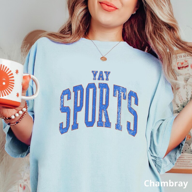 A woman wearing a light blue t-shirt with the text "YAY SPORTS" printed on it, holding a white mug with an orange design.