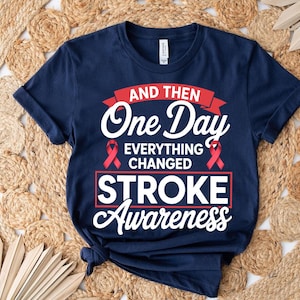 May include: Navy blue t-shirt with the text "AND THEN One Day EVERYTHING CHANGED STROKE Awareness" in white and red. The shirt features a crew neck and short sleeves. A red ribbon is displayed on the shirt.
