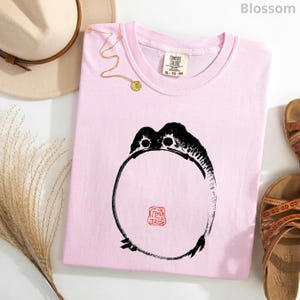 May include: A light pink t-shirt featuring a black ink-style circular frog illustration with a small red square stamp design in the center. The shirt has a Comfort Colors brand label at the inner neckline, size XL, and a crew neck collar.