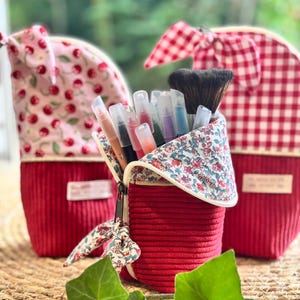 Peut inclure: Trois trousses en tissu verticales avec une base en velours côtelé rouge et des hauts à motifs. La trousse centrale possède un rabat à fleurs et contient divers stylos et un pinceau de maquillage. Les autres ont des motifs cerises et vichy.