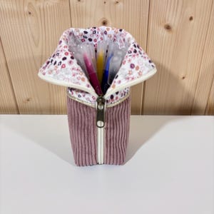 Op de afbeelding: Een mauve corduroy etui met een bloemenpatroon kraag en een witte rits. De etui is open en onthult verschillende kleurrijke pennen en stiften binnenin. De achtergrond is een licht houten paneel.