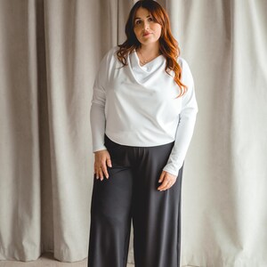 Peut inclure: Une femme porte un haut blanc à col bénitier et un pantalon noir à jambes larges. La tenue est simple et élégante, adaptée à diverses occasions. L'arrière-plan présente des rideaux de couleur neutre.