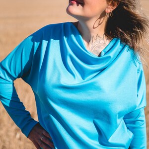 Peut inclure: Un haut turquoise à manches longues avec un col bénitier. Le haut est fait d'une matière douce et est associé à un bas noir. L'arrière-plan présente un champ d'herbe sèche sous un ciel bleu vif.