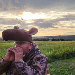 May include: A person wearing a brown moose hat and camouflage jacket stands in a field. The hat has large ears. The background features a green field, a cloudy sky, and a silo.